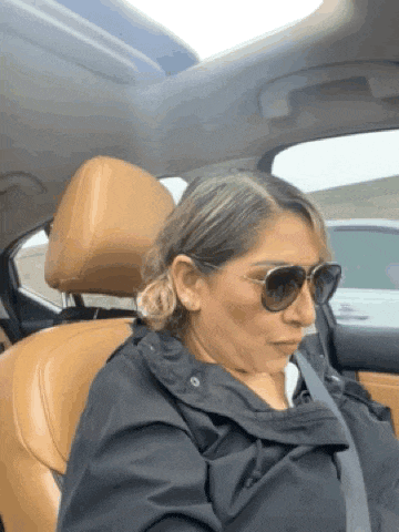 a woman wearing sunglasses and a black jacket is sitting in the driver 's seat of a car .