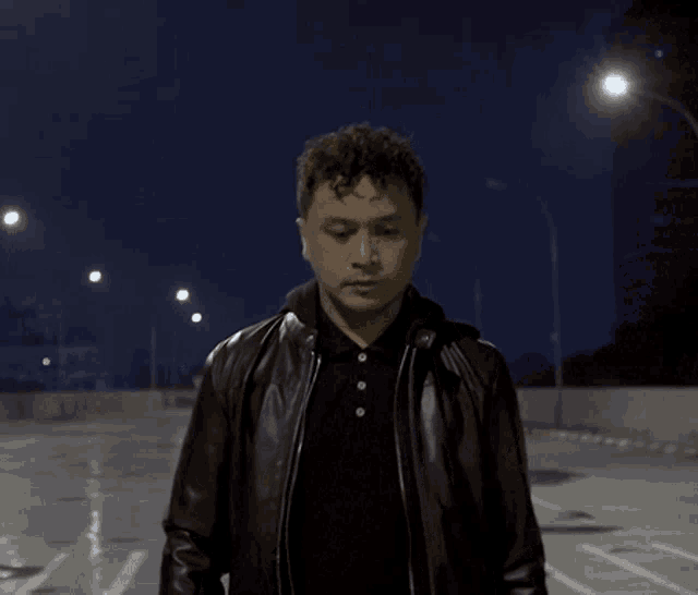 a man in a leather jacket and black shirt stands in a parking lot at night