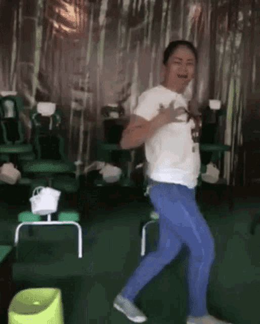 a woman in a white shirt and blue jeans is dancing in a room with green chairs