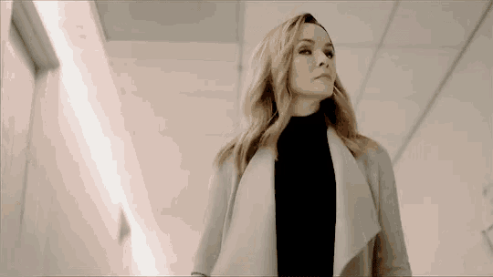 a woman in a white coat and black turtleneck is standing in a hallway looking at the camera .