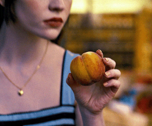 a woman is holding a peach in her hand and wearing a necklace with a heart on it