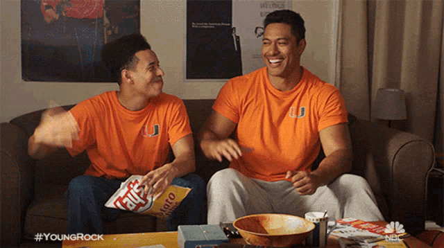 two men are sitting on a couch with a bag of potato chips in front of them