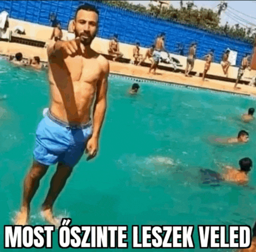 a shirtless man in blue shorts is standing in a swimming pool with people swimming in the background .