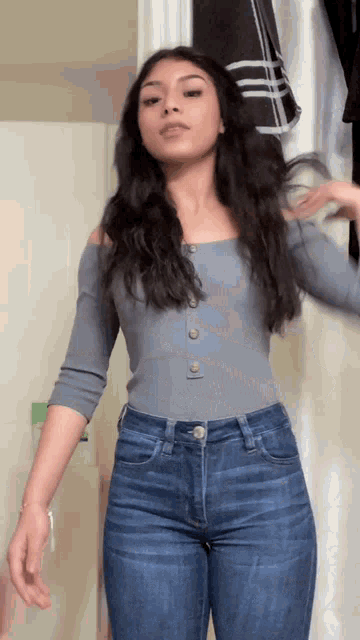 a woman wearing a grey off the shoulder top and blue jeans is standing in front of a shower curtain .