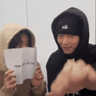 a man in a hoodie is holding a piece of paper in front of his face while another man looks on .