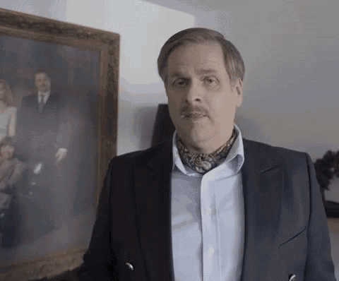 a man in a suit and tie with a mustache is standing in front of a painting of a family .