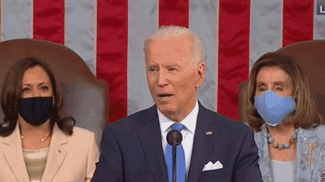 a man in a suit and tie is speaking into a microphone in front of two women wearing masks .