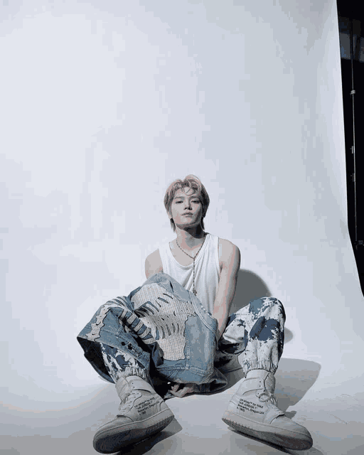 a man with pink hair sits on the floor wearing a white tank top and blue jeans