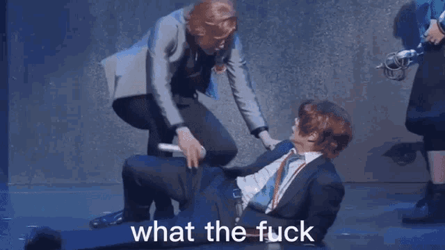 a man in a suit and tie is laying on the floor while another man stands behind him and says what the fuck