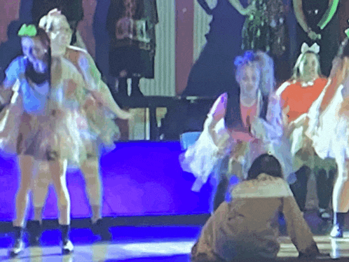 a group of young girls are dancing on a stage with a man sitting on the floor