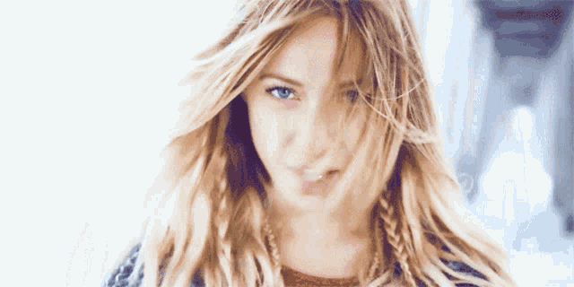 a woman with blonde hair and blue eyes looks at the camera with her hair blowing in the wind