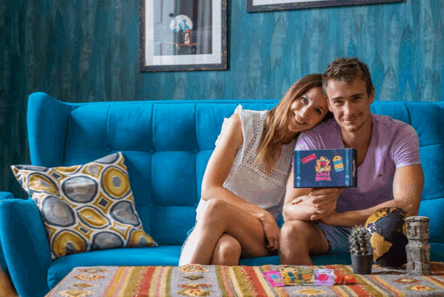 a man and woman are sitting on a blue couch holding a box that says ' happy birthday '