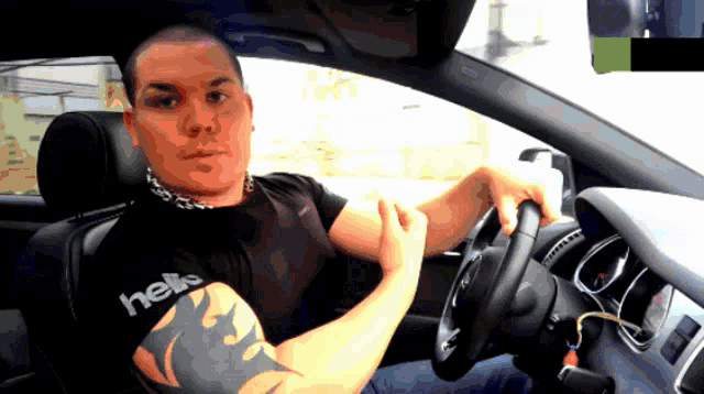 a man in a black shirt with the word hells on it is driving a car