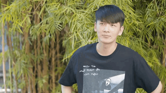 a young man wearing a black t-shirt that says " meow this " stands in front of a bamboo tree