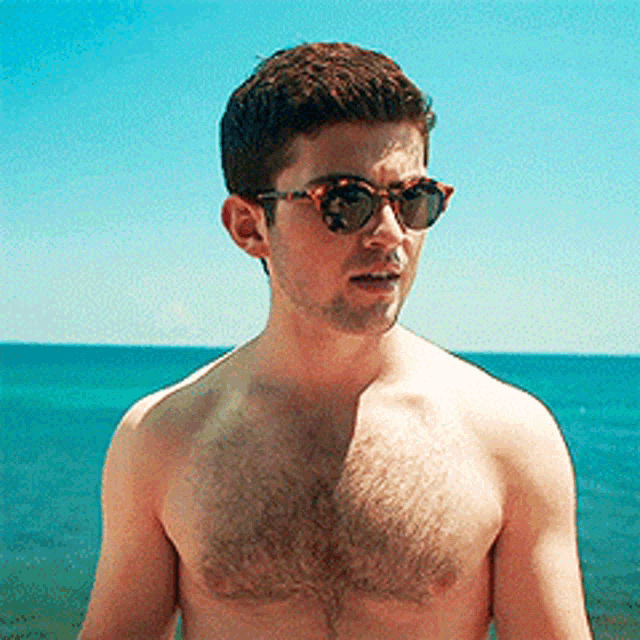 a shirtless man wearing sunglasses stands in front of the ocean