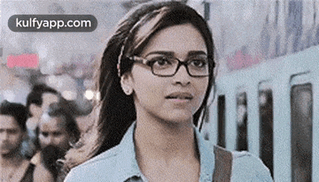 a woman wearing glasses is standing in front of a train and looking at the camera .