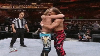 two men are hugging each other in a wrestling ring while a referee looks on .