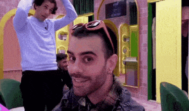 a man wearing sunglasses looks at the camera with another man behind him