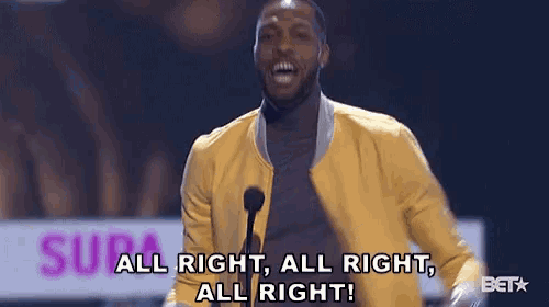 a man in a yellow jacket stands in front of a microphone and says all right all right all right