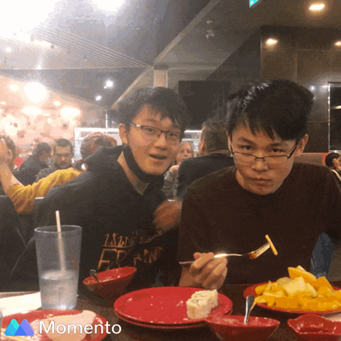 two young men are sitting at a table with plates of food and a momento logo