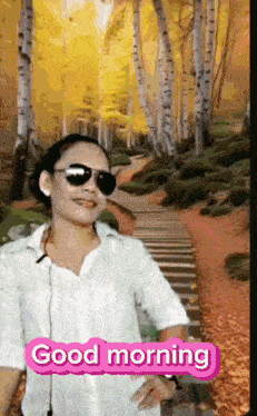 a woman wearing sunglasses and a white shirt is standing in front of a forest and says good morning