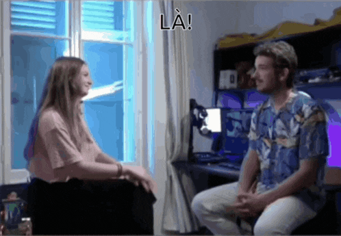 a man and a woman are sitting in front of a window with la written on the bottom right
