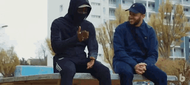 two men are sitting on a bench and talking to each other . one of the men is wearing a mask .