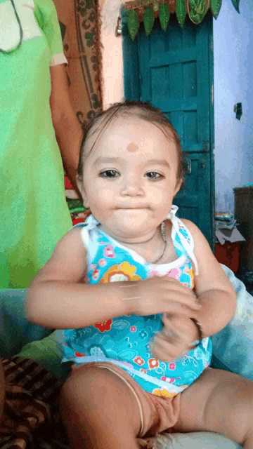 a baby wearing a blue tank top with flowers on it looks at the camera