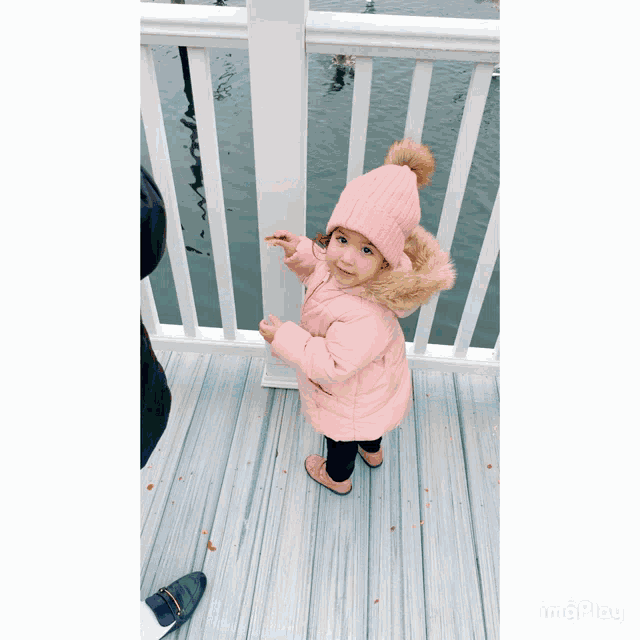 a little girl wearing a pink hat and a pink coat stands on a deck