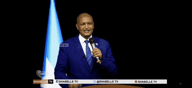 a man in a blue suit and tie is speaking into a microphone in front of a flag