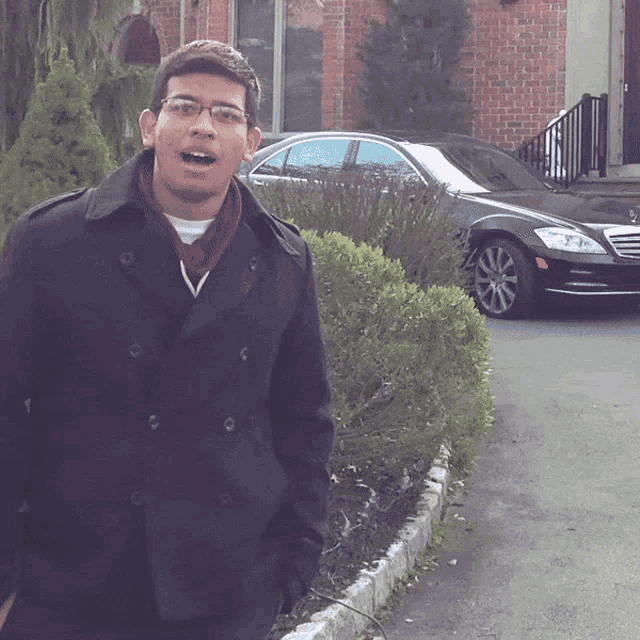 a man wearing glasses and a black coat stands in front of a black car