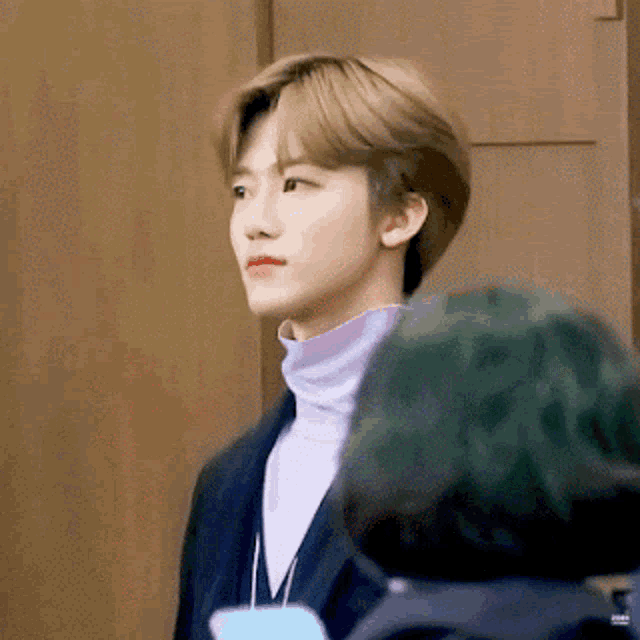 a young man wearing a turtleneck sweater and a jacket is standing in front of a wooden wall .