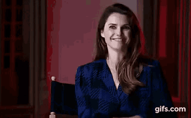 a woman in a blue jacket is sitting in a chair in front of a red curtain and smiling .
