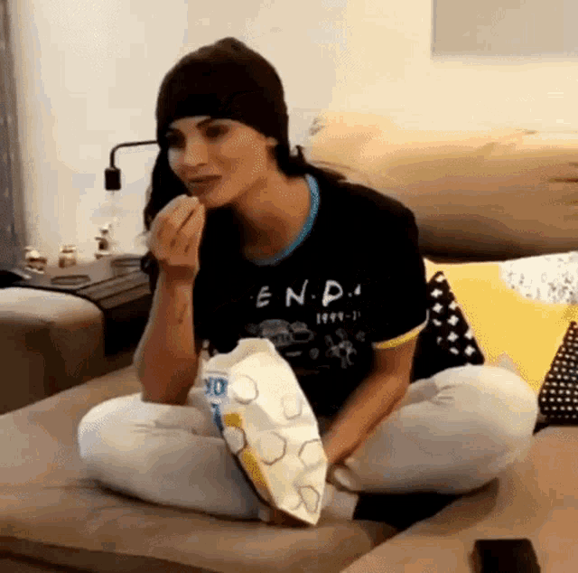 a woman is sitting on a couch eating popcorn and wearing a beanie .