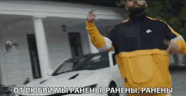 a man in a yellow and black jacket is standing in front of a white car and giving a middle finger .