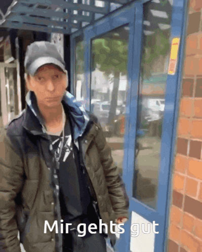 a man wearing a hat and a jacket is standing in front of a blue door with the words mir gehts gut written on it