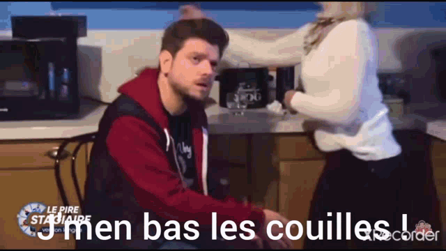 a man is sitting in a kitchen with a woman standing behind him and the words men bas les couilles on the bottom