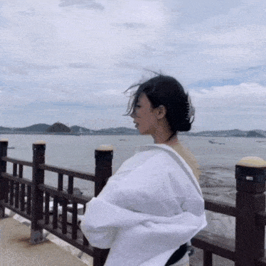 a woman in a white shirt stands in front of a wooden railing overlooking the ocean