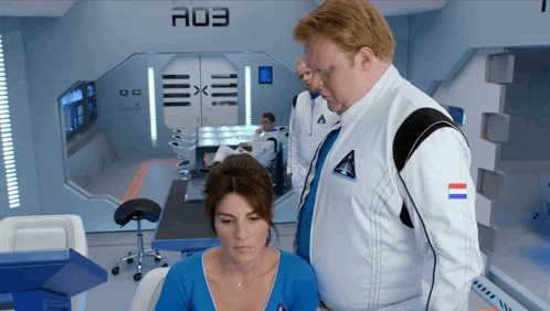 a man in a space suit stands next to a woman in a room with the number a03 on the wall behind them