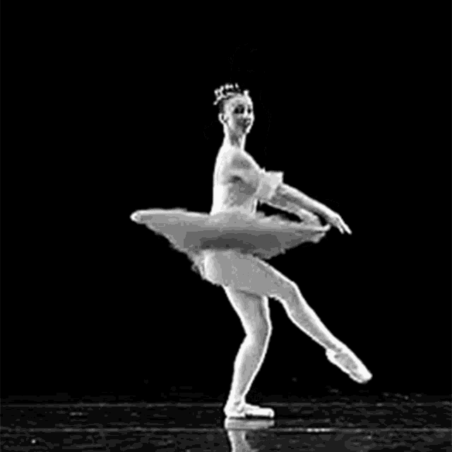 a black and white photo of a ballerina in a tutu standing on one leg on a black background .