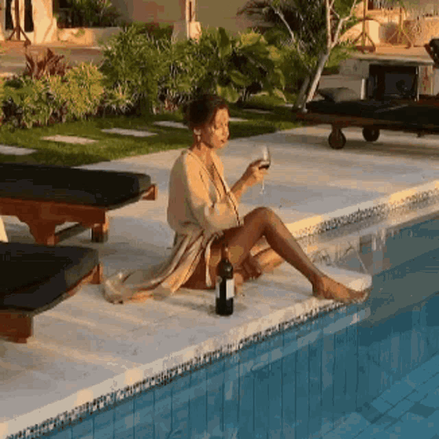 a woman sits on the edge of a swimming pool with a bottle of wine