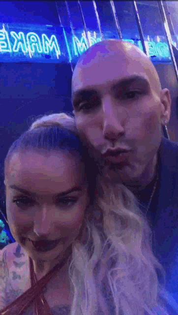 a bald man and a blonde woman are posing for a picture in front of a neon sign that says exam