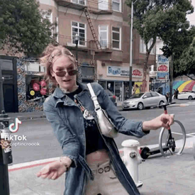 a woman wearing sunglasses and a denim jacket is standing in front of a fire hydrant on a street .