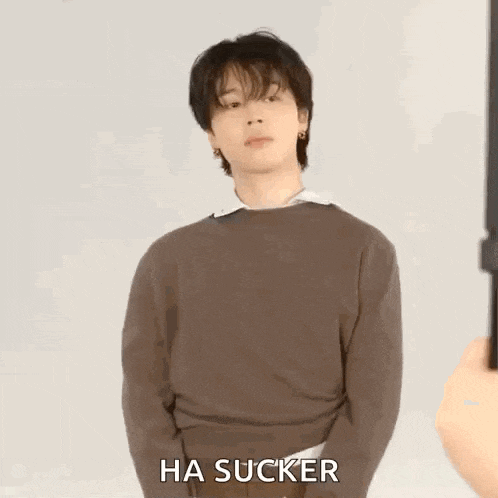 a young man in a brown sweater is standing in front of a white wall and making a funny face .