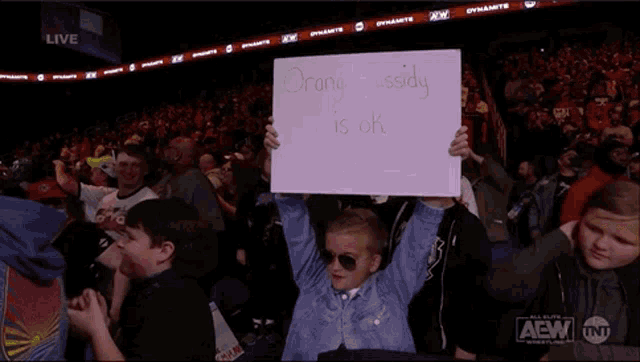 a young boy holds up a sign that reads orange cassidy is ok