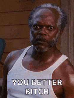 a man with a beard and a white tank top is standing in front of a building and says `` you better bitch '' .