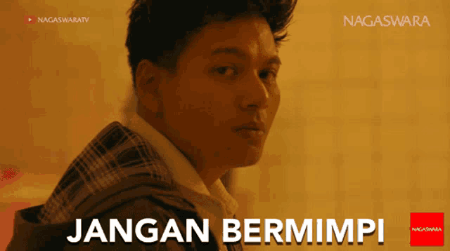 a man in a plaid jacket is standing in front of a sign that says " jangan bermimpi "