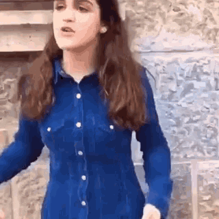 a woman wearing a blue shirt is standing in front of a stone wall .