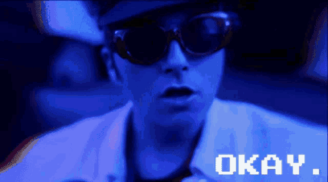 a man wearing sunglasses and a hat says okay on a blue screen