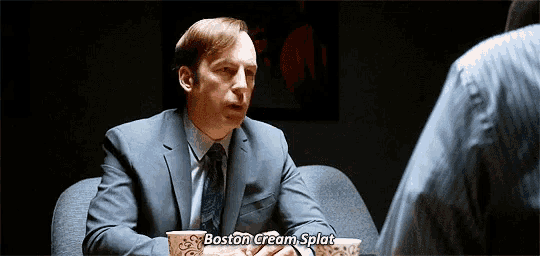 a man in a suit and tie is sitting at a table with a cup of boston cream splat .